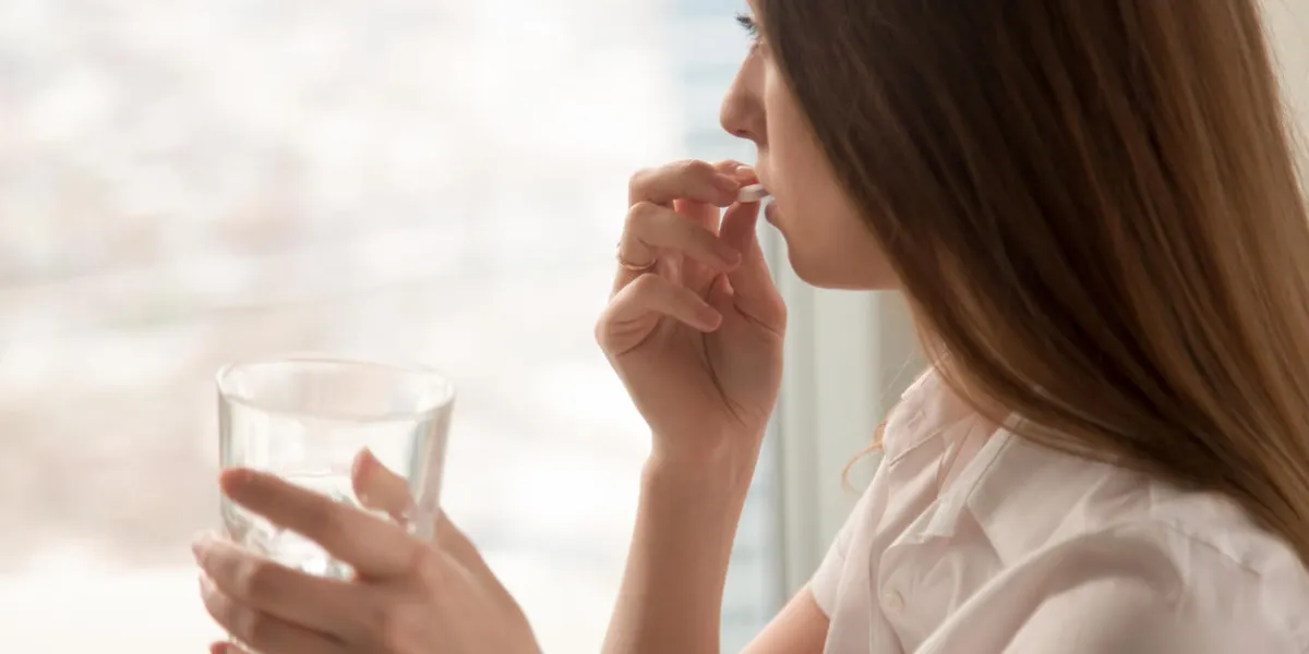 young woman takes white round pill with glass of water in hand stressed female looking in window and drinking sedated antidepressant meds woman feels depressed, taking drugs medicines at work