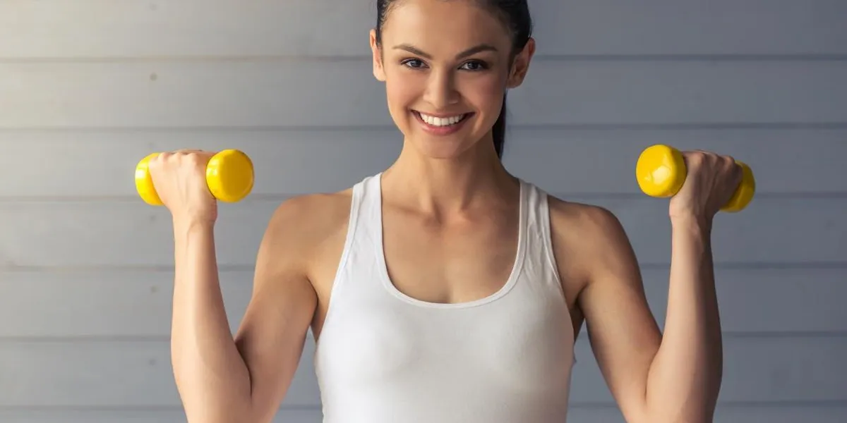 beautiful young woman in sports wear is working out with dumbbells, looking at camera and smiling, standing on a gray background