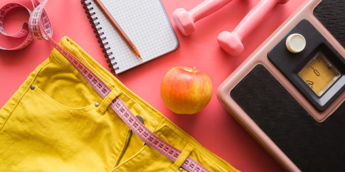 measure tape with yellow jeans, dumbbells, weight scale, apple, notebook on a pink background women diet before summer season healthy lifestyle, body slimming, weight loss concept cares about body