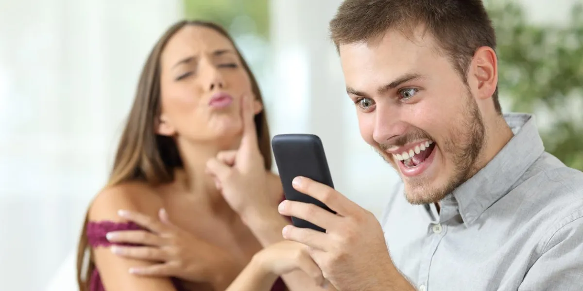 man with a smart phone ignoring a sex proposal of his girlfriend sitting on a sofa at home