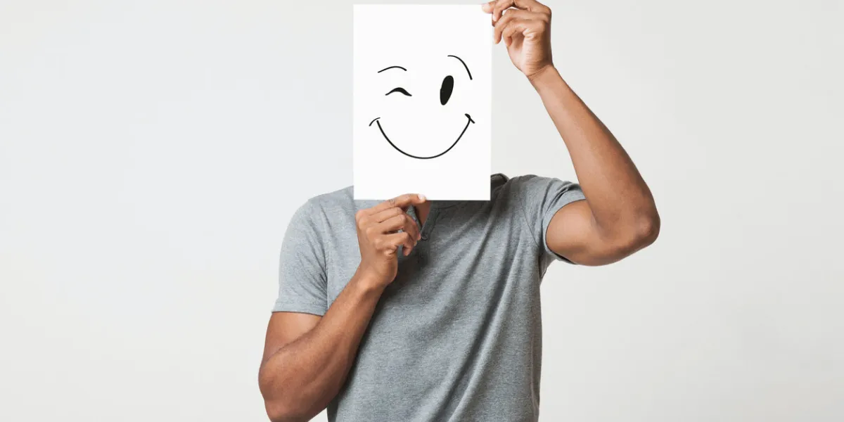 black man cover his face with happy and winking smiley drawn on paper, white studio background