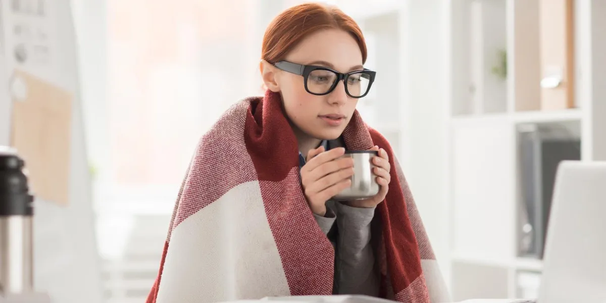 young woman in plaid and eyeglasses drinking hot tea from mug during work in office