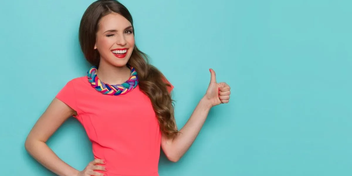 smiling beautiful young woman in pink mini dress showing thumb up and winking three quarter length studio shot on turquoise background