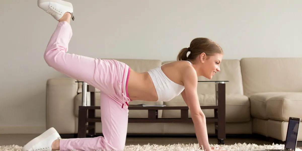 pretty young lady in sportswear doing one knee kickbacks and watching aerobics videos on laptop, repeating exercise with online personal fitness trainer while training on carpet in living room at home pretty young lady in sportswear doing one knee kickbacks and watching aerobics videos on laptop, repeating exercise with online personal fitness trainer while training on carpet in living room at home