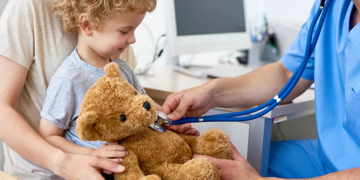 portrait of adorable curly child sitting on mothers lap in doctors office holding teddy bear toy, with pediatrician listening to heartbeat using stethoscope