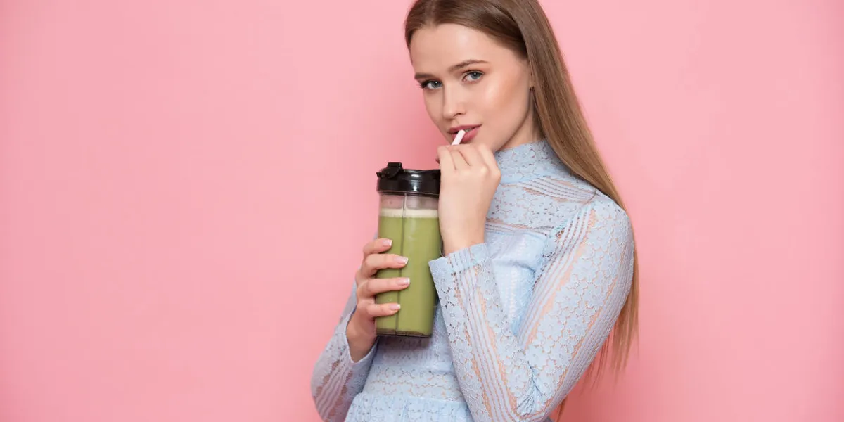 beautiful young adult woman drinking healthy green smoothie cocktail near pink wall