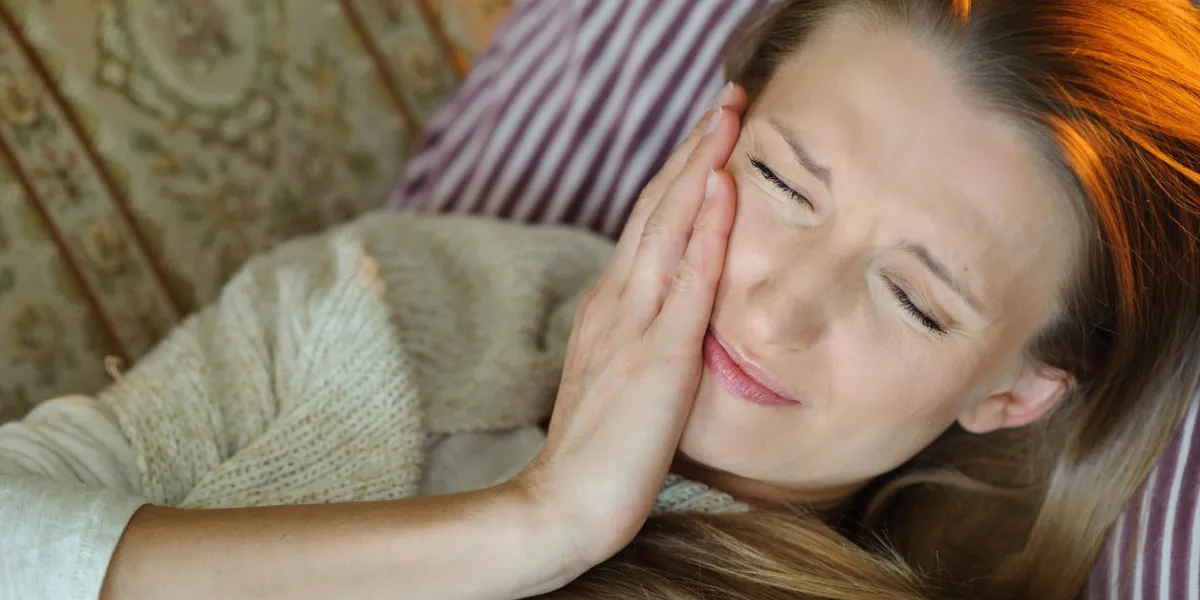 woman with toothache and pain lying on sofa at home with hand on cheek