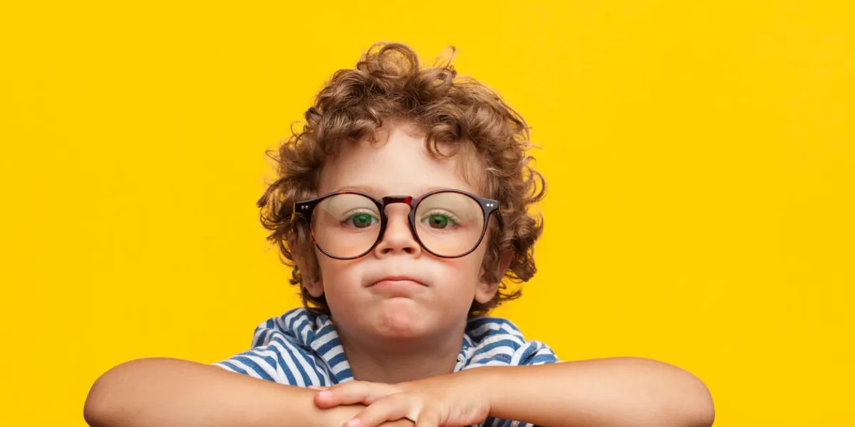 little curly boy in glasses leaning on wooden box looking at camera on orange background