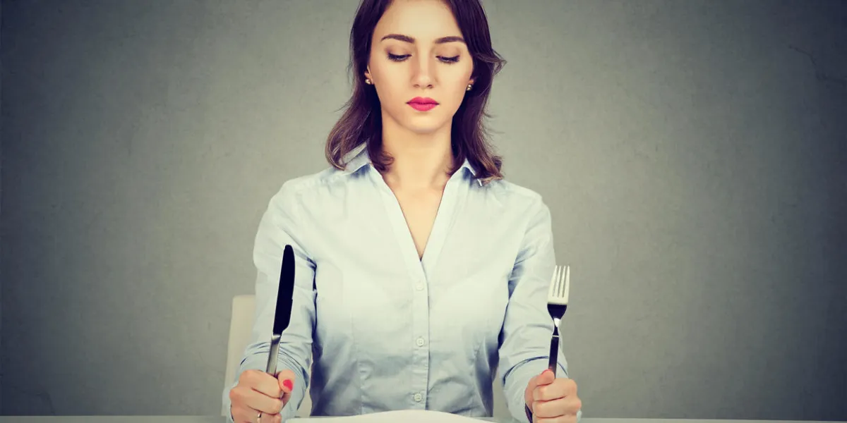 serious woman with fork and knife sitting at table with empty plate isolated on gray wall background serious woman with fork and knife sitting at table with empty plate isolated on gray wall background