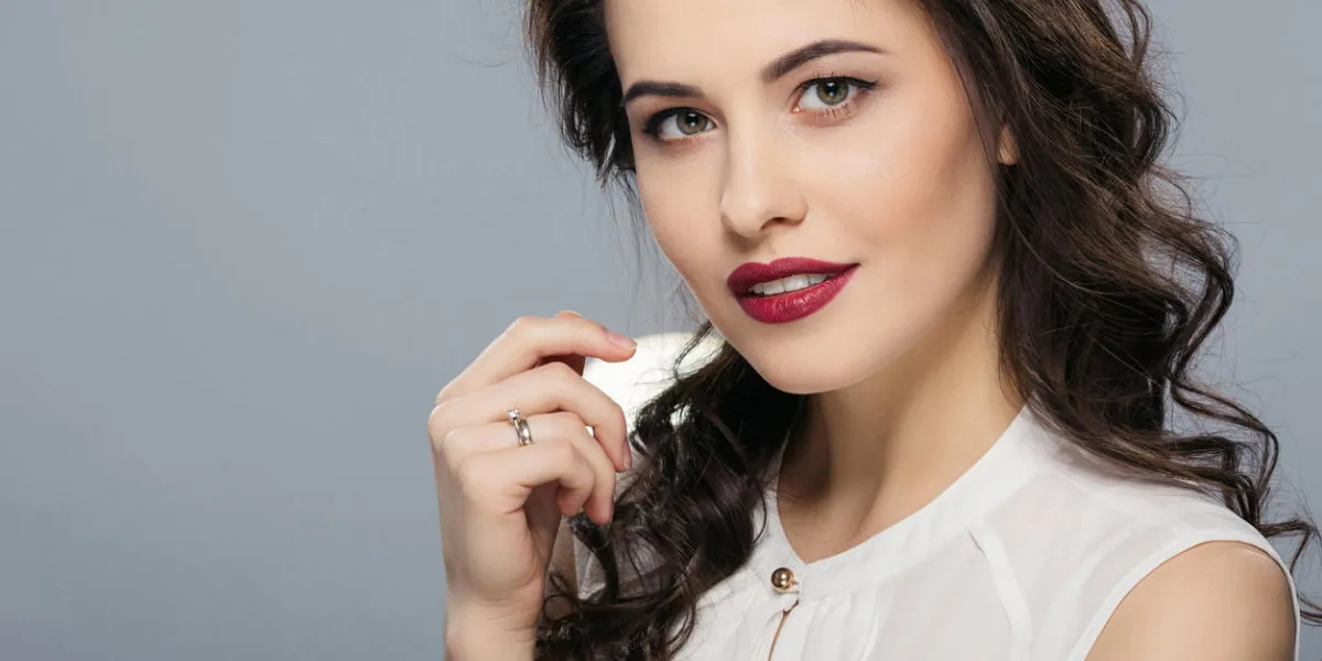 femme aux cheveux longs bouclés, grands yeux et lèvres rouges portant chemise blanche regardant la caméra, tenant la main près de face, fond de studio gris, espace copie