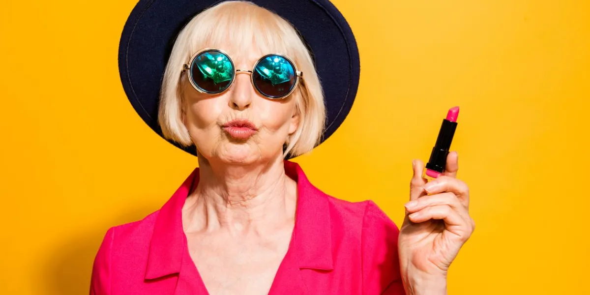 grandmother getting ready for date charming, attractive old woman holding red lipstick pouting lips for a kiss isolated on brigth yellow background