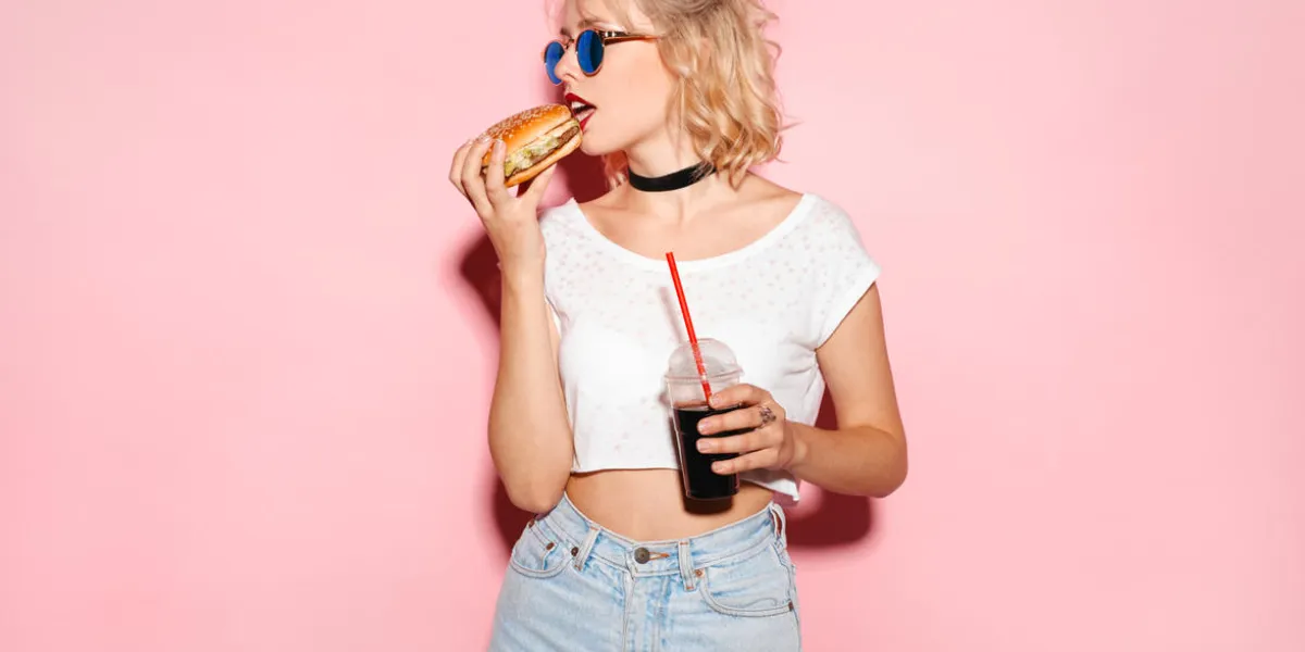 young woman holding a glass of coke and biting a burger horizontal studio shot young woman holding a glass of coke and biting a burger horizontal studio shot