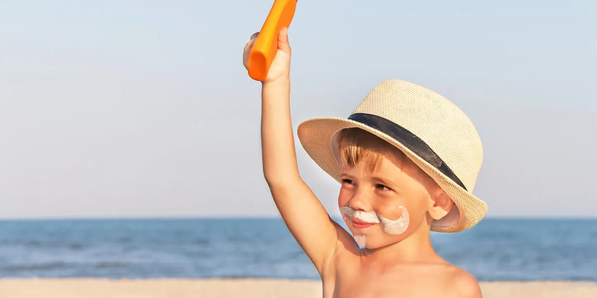 the mustache drawing sunscreen (suntan lotion) on hipster baby (boy) face caucasian smiling, funny child (kid) is holding plastic container of sunscreen on sunny beach trendy fashion concept copy space, close up, outdoor