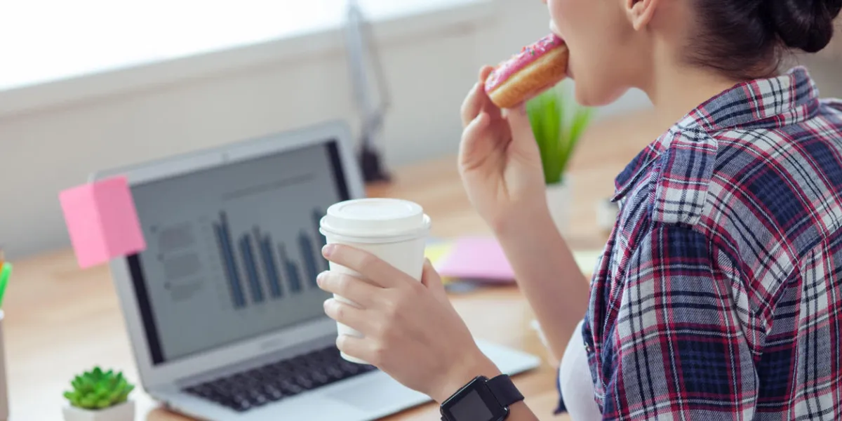 cheerful young woman is making break after work she is eating a donut and drinking coffee with pleasure the lady is sitting at desk near the laptop