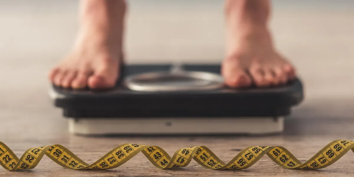 cropped image of woman feet standing on weigh scales, on gray background a tape measure in the foreground