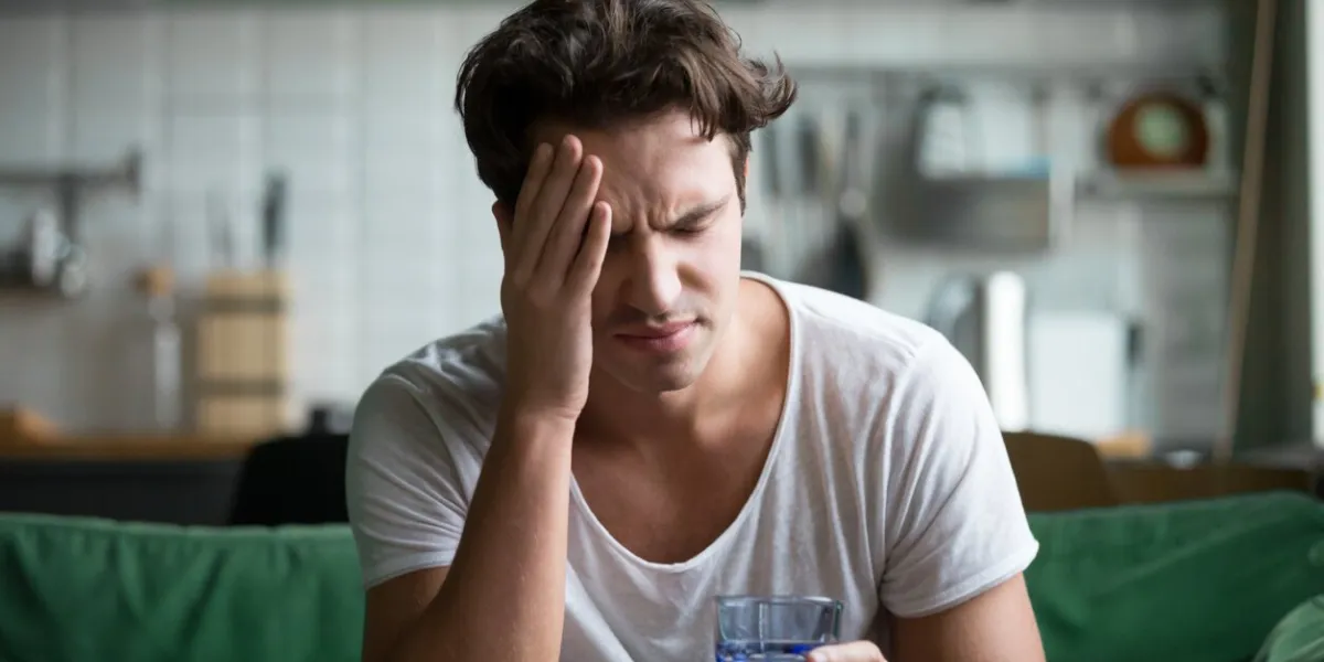 young man suffering from strong headache or migraine sitting with glass of water in the kitchen, millennial guy feeling intoxication and pain touching aching head, morning after hangover concept young man suffering from strong headache or migraine sitting with glass of water in the kitchen, millennial guy feeling intoxication and pain touching aching head, morning after hangover concept