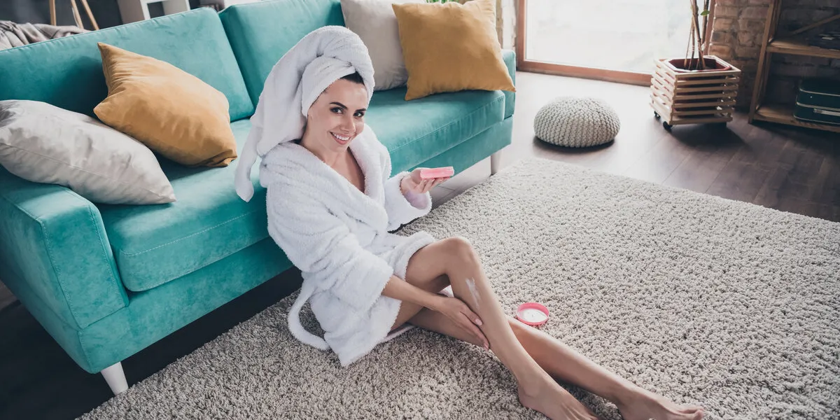 portrait of attractive lady apply body cream lotion balm after legs waxing epilation sitting floor carpet spacious flat living room indoors
