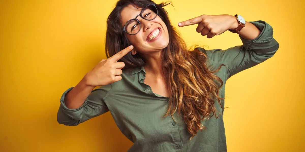 young beautiful woman wearing green shirt and glasses over yelllow isolated background smiling cheerful showing and pointing with fingers teeth and mouth dental health concept young beautiful woman wearing green shirt and glasses over yelllow isolated background smiling cheerful showing and pointing with fingers teeth and mouth dental health concept