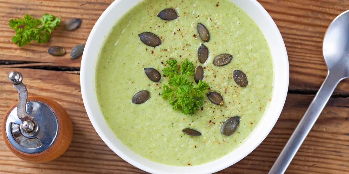 photographié du haut, un bol blanc de soupe de crème de brocoli fraîchement saupoudré de graines de citrouille et de persil servi sur une vieille table en bois fissuré avec un moulin à poivre rustique