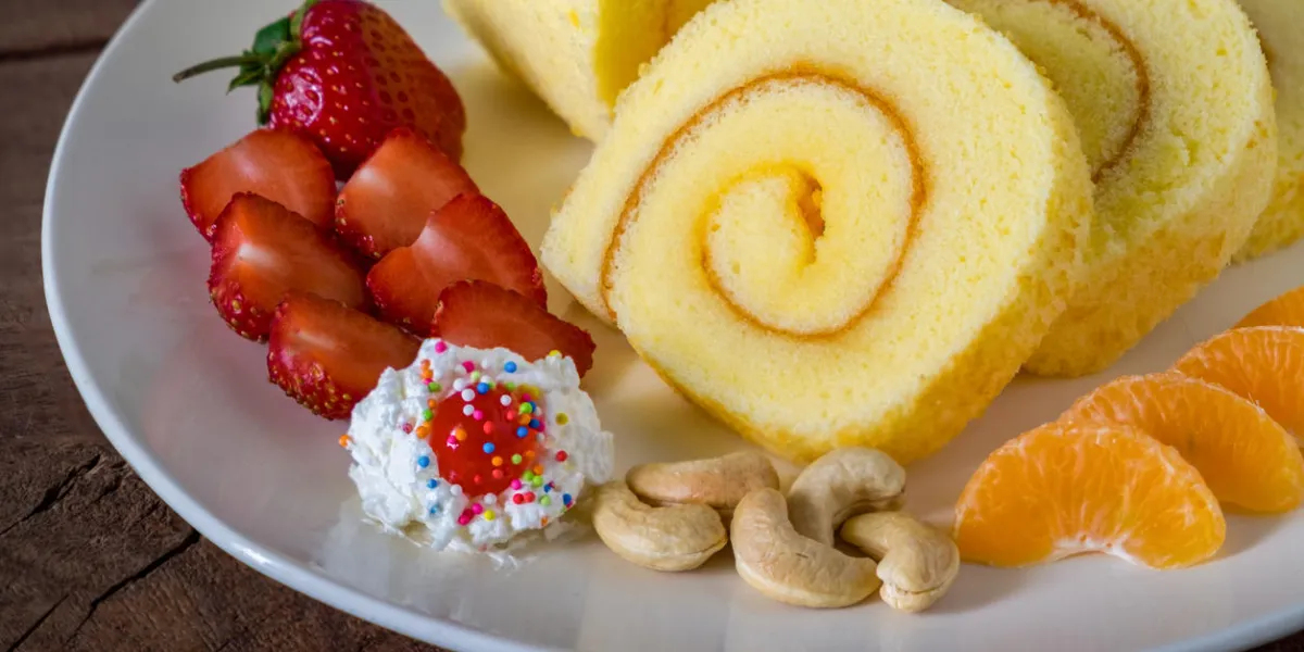 sponge cake roll with cream, strawberries and orange fruits on a white plate on wooded table background sponge cake roll with cream, strawberries and orange fruits on a white plate on wooded table background