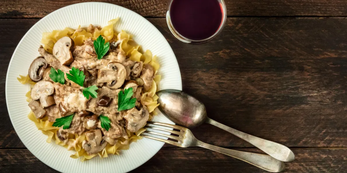 mushroom beef stroganoff, served with the typical egg pasta, with cremini and champignons, with a glass of red wine, a fork, and a spoon, shot from above on a dark rustic texture with a place for text mushroom beef stroganoff, served with the typical egg pasta, with cremini and champignons, with a glass of red wine, a fork, and a spoon, shot from above on a dark rustic texture with a place for text
