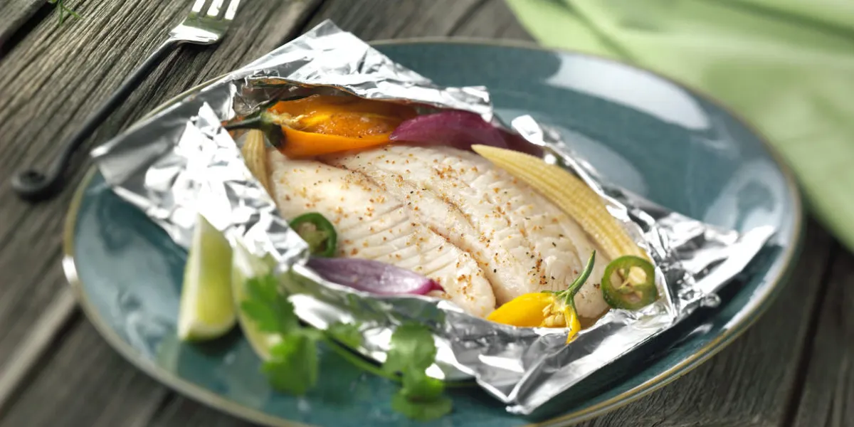 tilapia fish cooked in foil with vegetables on plate on wood background tilapia fish cooked in foil with vegetables on plate on wood background